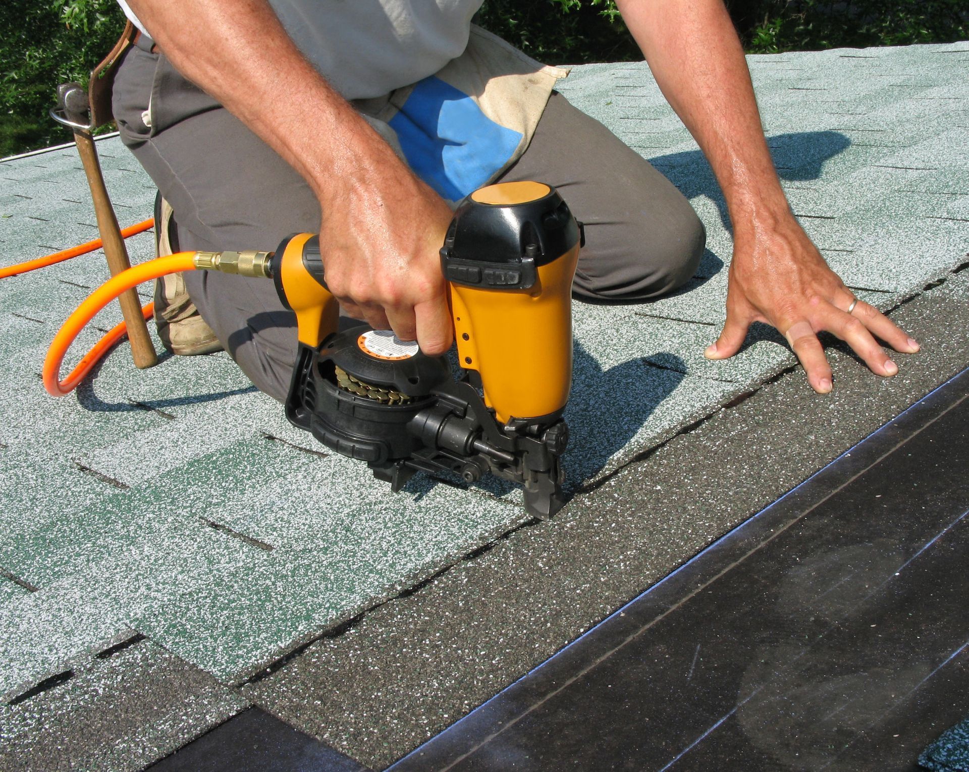 A person kneeling on a roof uses a nail gun to attach roofing material.