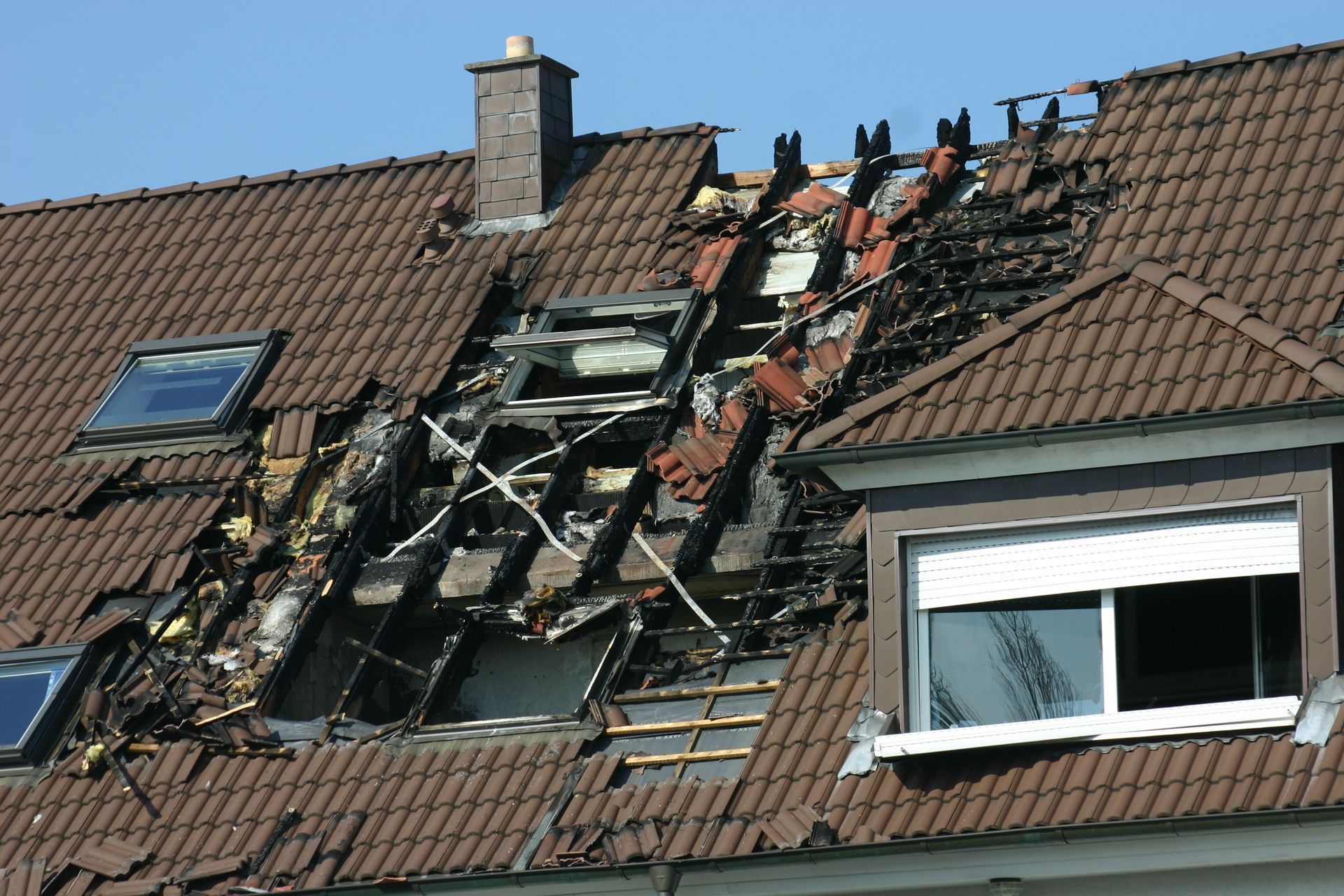 Burnt roof with missing tiles, exposing charred wooden beams and skylights, against a blue sky.
