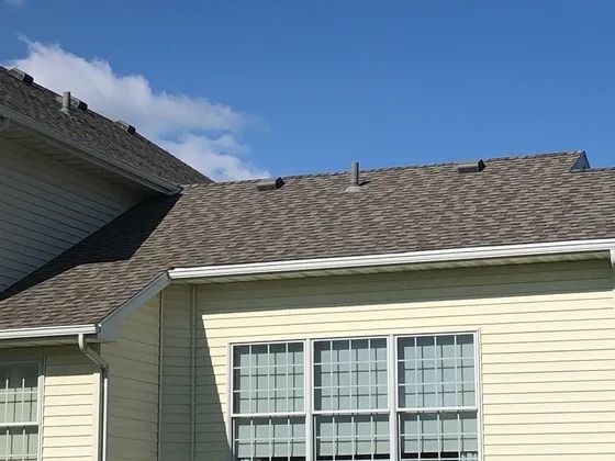Roof of a house with gray shingles, light yellow siding, white gutters, and blue sky with clouds.