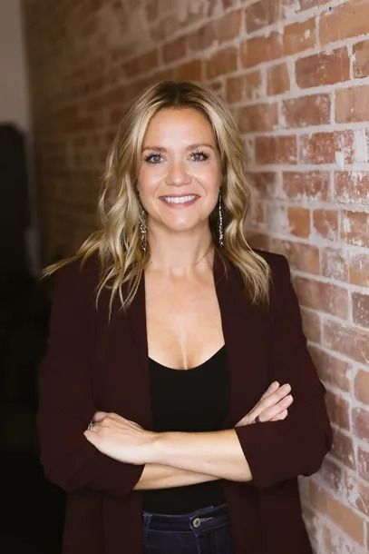 Woman with blonde hair, arms crossed, smiles at camera. Leaning against a brick wall, wearing a dark jacket.