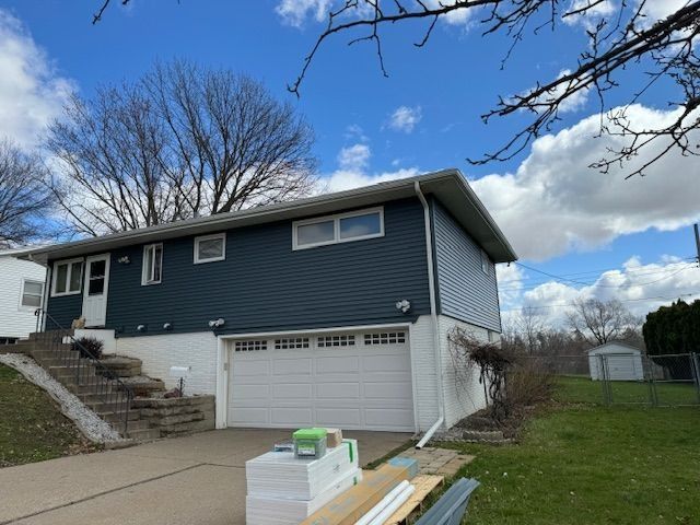 Blue-sided two-story house with white garage and trim, cloudy blue sky, work materials in foreground.