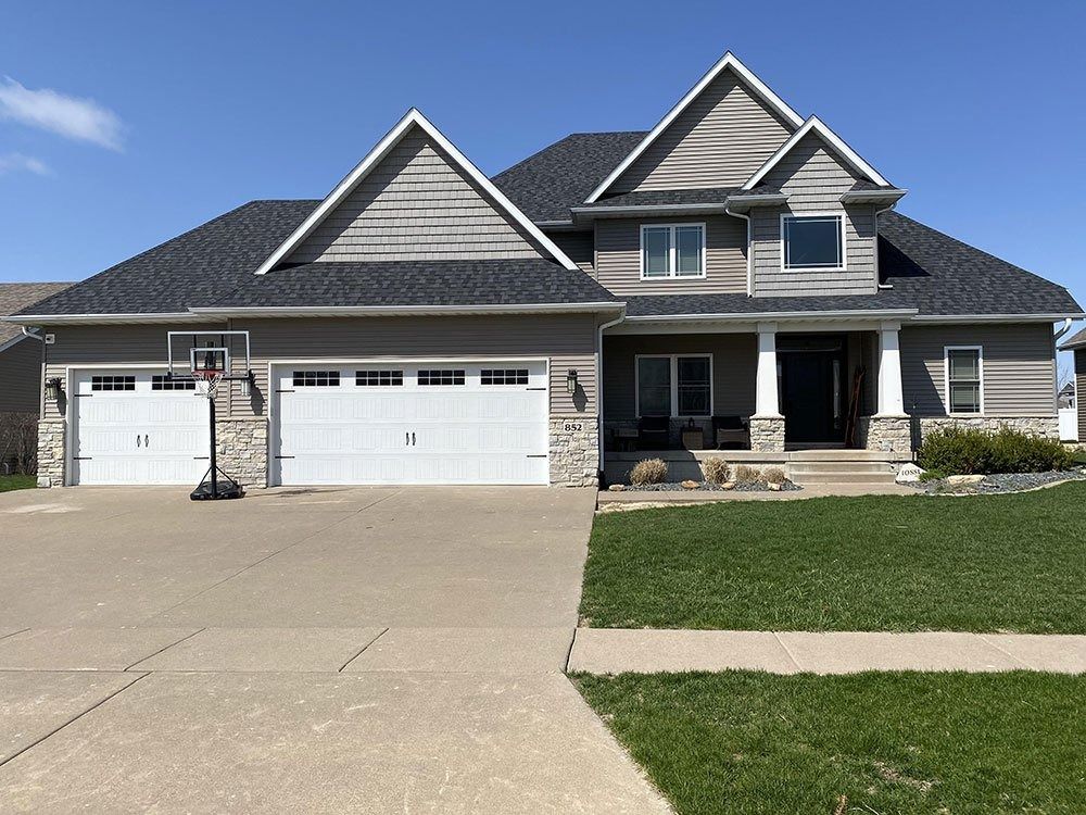 Gray two-story house with white garage doors and columns, stone accents, and a basketball hoop in front of a blue sky.