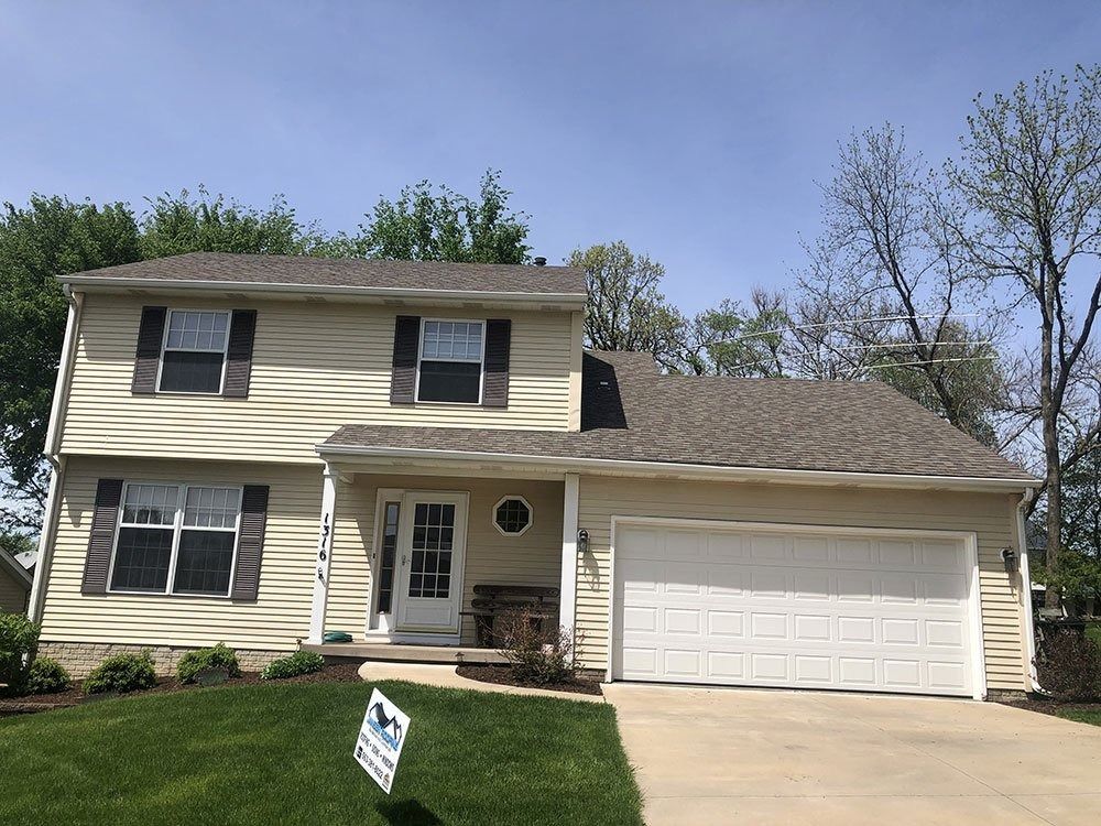 Two-story beige house with a brown roof and garage. Green lawn with trees and a blue sky in the background.