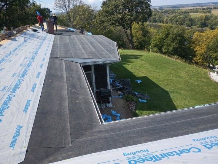 Workers on a roof installing roofing material. Green lawn and trees in the background.