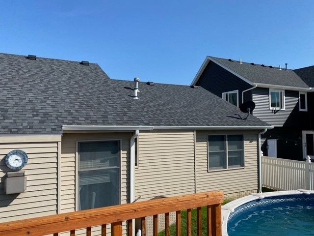 Backyard with pool, tan siding, dark shingle roof, blue sky. Adjacent house with gray siding.