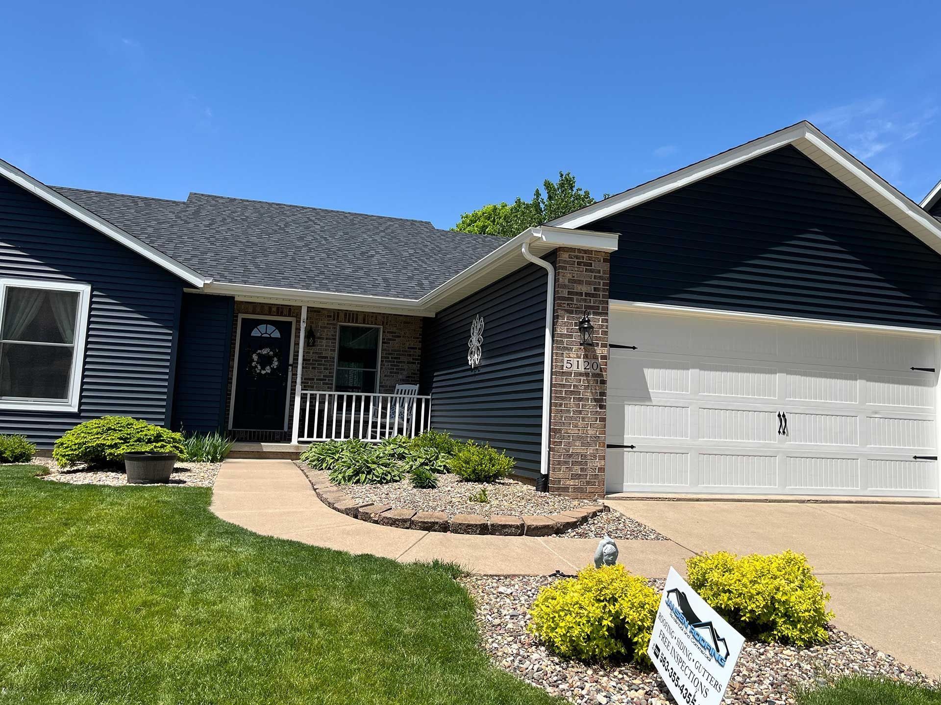 Blue house with a black roof, stone column, and white garage door. Landscaped front yard with green grass.