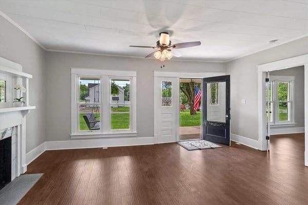 Bright empty living room with hardwood floors, white walls, ceiling fan, fireplace, and open front door.