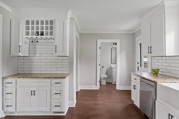 Bright white modern kitchen with island, subway tile backsplash, and hardwood floors leading to a bathroom.