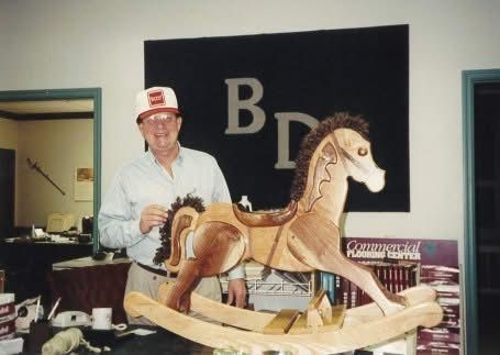 Man standing beside a wooden rocking horse display in an indoor room with a “BD” sign on the wall
