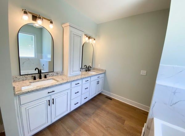 Bright bathroom vanity with double sinks, arched mirrors, white cabinets, and wood-look flooring