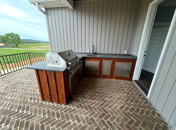 Covered patio with outdoor kitchen, grill, cabinets, and brick herringbone floor overlooking a grassy field