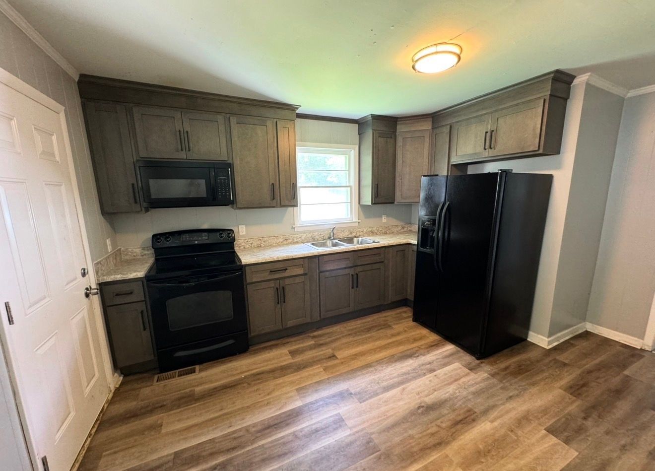Small kitchen with dark cabinets, black appliances, wood floor, and a window over the sink.