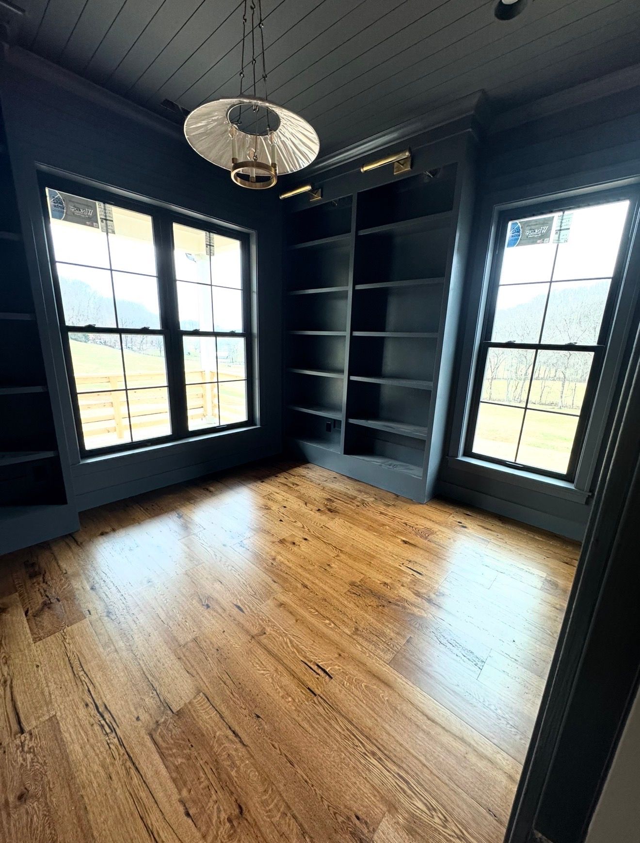 Empty dark-blue room with hardwood floor, tall windows, built-in shelves, and a hanging light fixture.