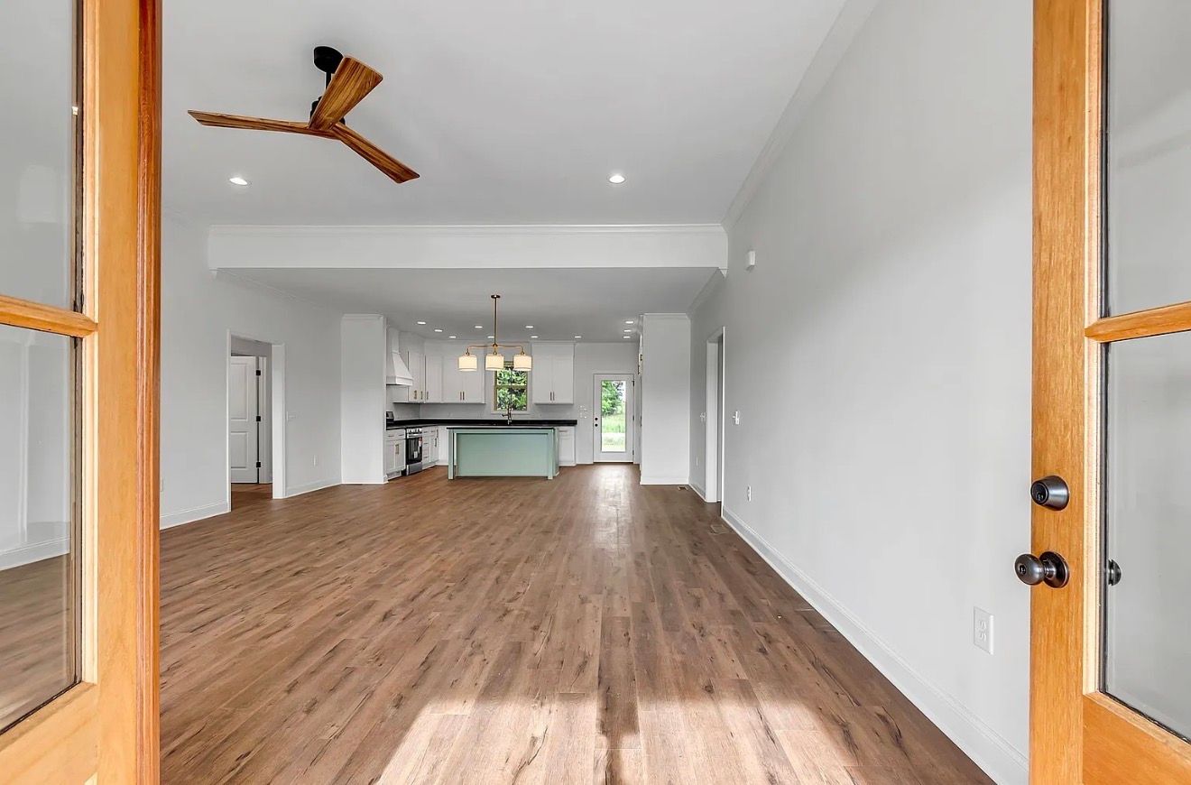 Empty modern living room with hardwood floors, white walls, and open kitchen view through French doors