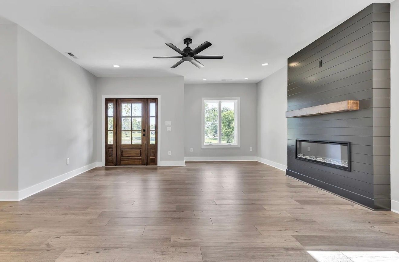 Empty modern living room with gray walls, wood floors, ceiling fan, and brick fireplace.