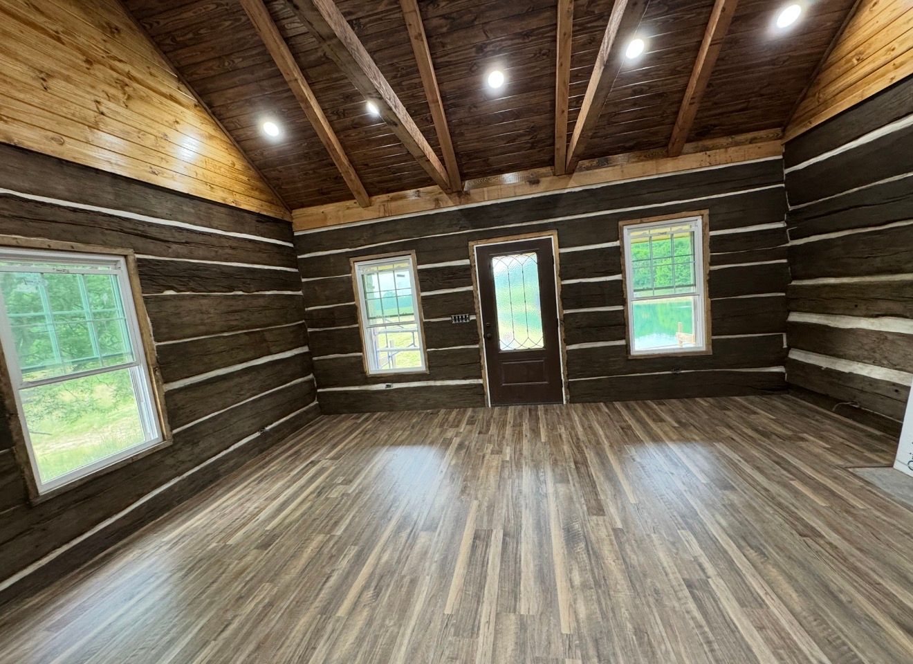 Empty rustic room with wood plank walls, exposed beams, and three windows plus a door.
