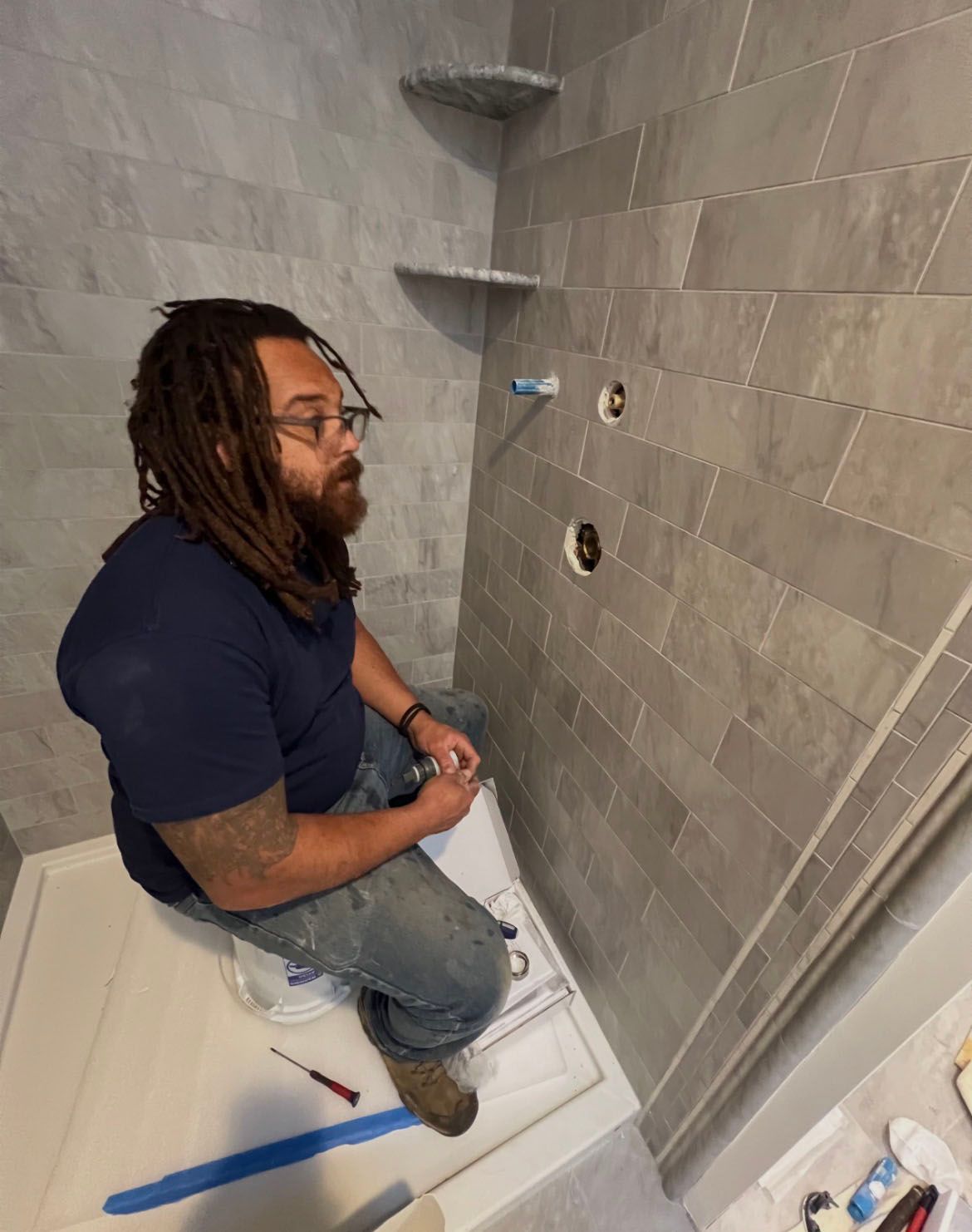 Person kneels in a partially tiled shower, holding tools. Plumbing fixtures visible on the wall.