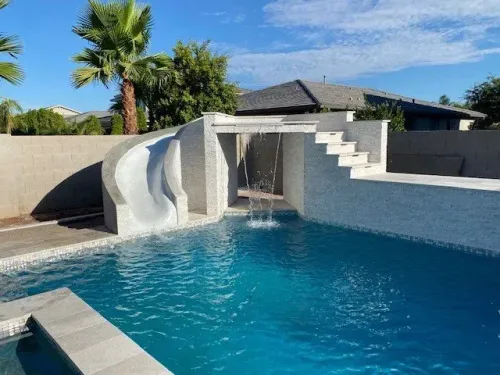 Pool with slide, waterfall, and steps against a white tiled structure, palm trees, and blue sky.
