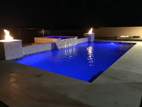 Nighttime view of a pool with blue underwater lighting, a waterfall, fire features, and a hot tub.