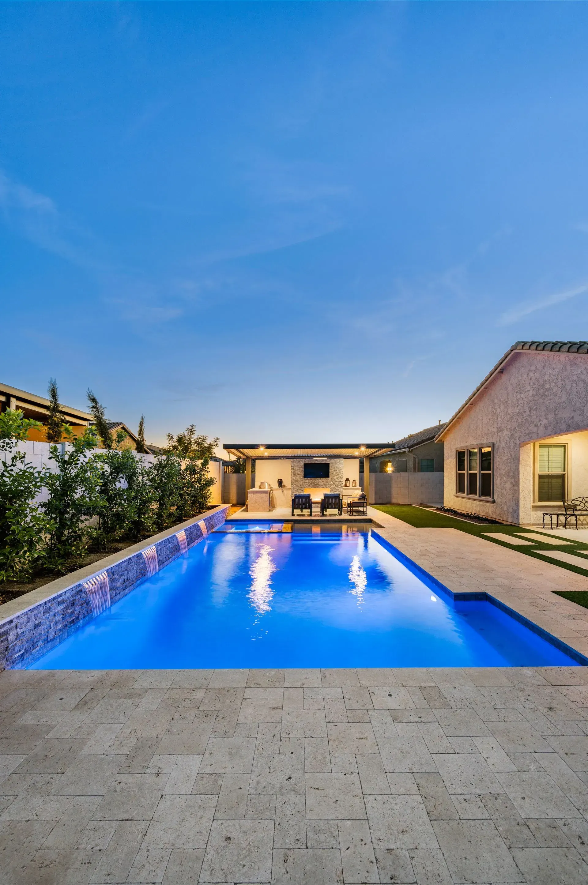 A rectangular pool with blue water and lights is flanked by a patio and a building under a dusk sky.