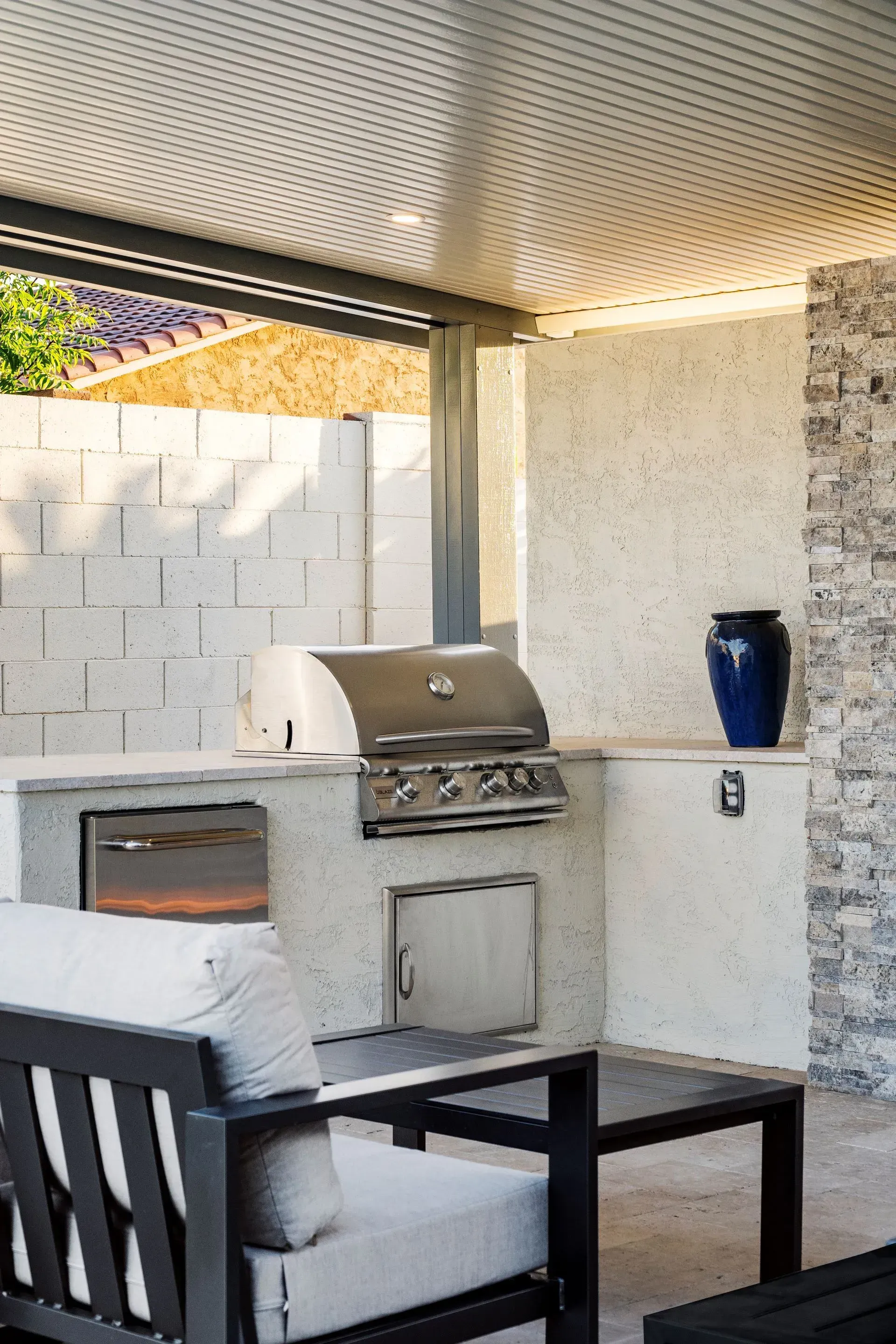 Outdoor kitchen with built-in grill, oven, and storage. Gray and white speckled countertops, brick wall, blue vase.