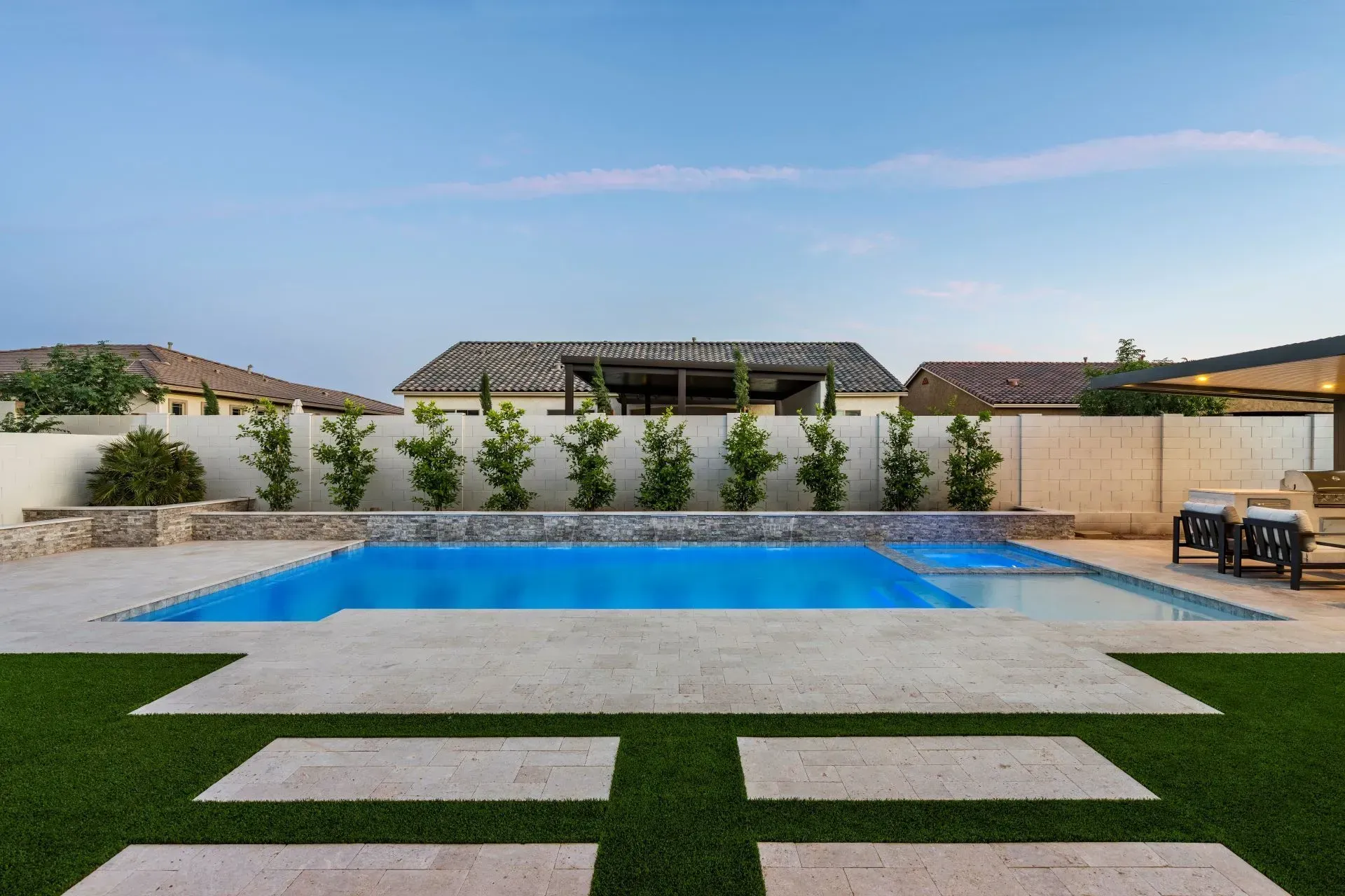 Pool area with blue water, stone patio, green grass, and white wall with trees.