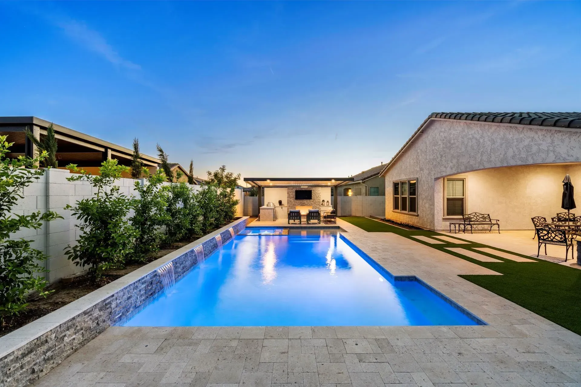 Backyard pool with a waterfall feature, and patio seating, at dusk.