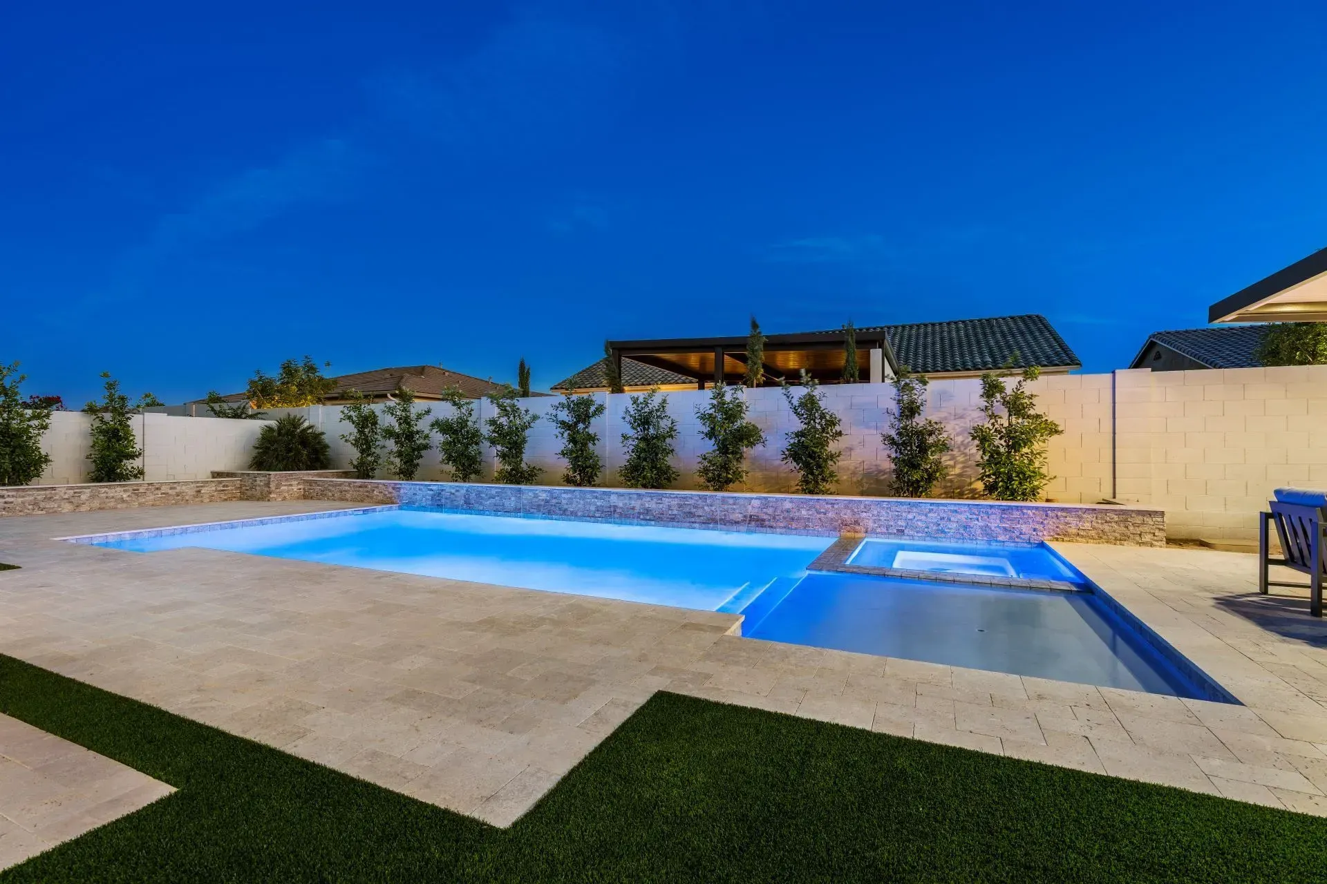 Backyard pool and spa illuminated at night with a blue glow; surrounded by a stone patio and landscaping.
