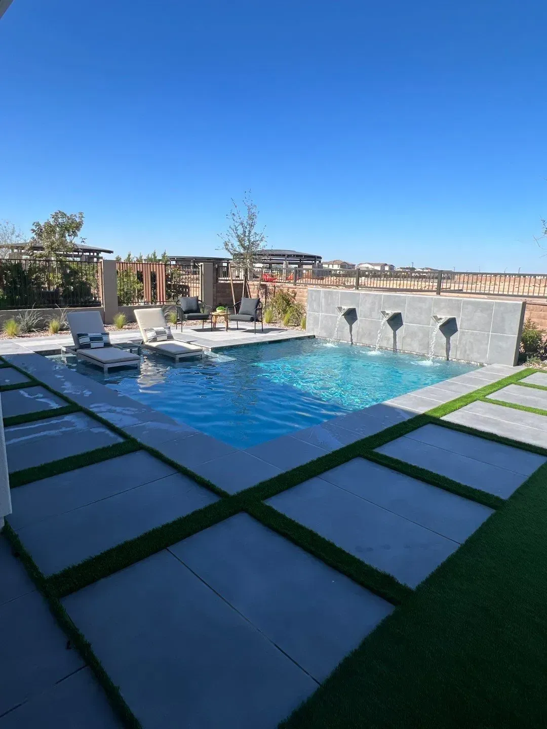 A pool with fountains and surrounding stone tiles with grass insets, under a clear blue sky.