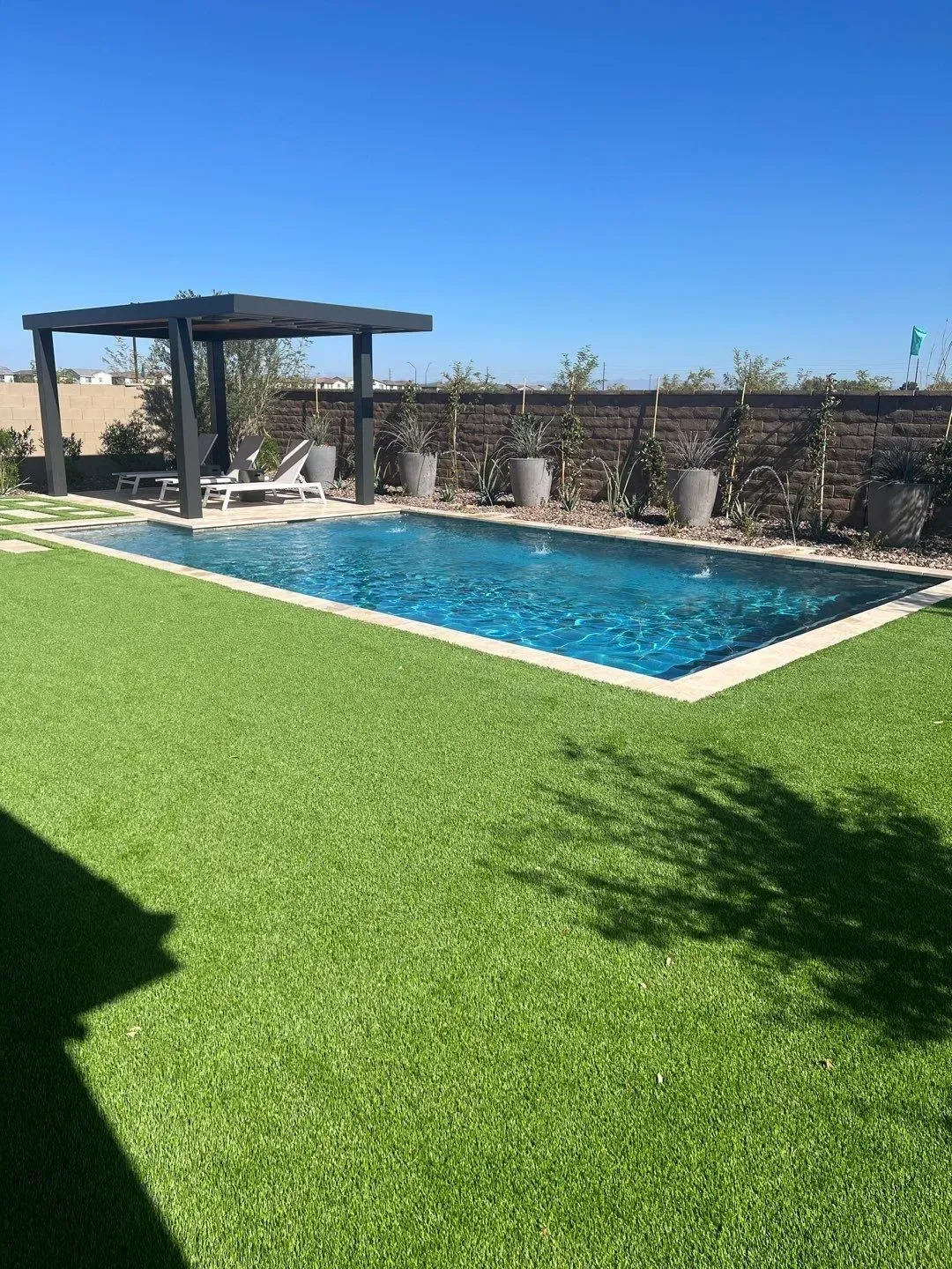 Rectangular pool with artificial green lawn, dark pergola, and potted plants against a brick wall under a blue sky.