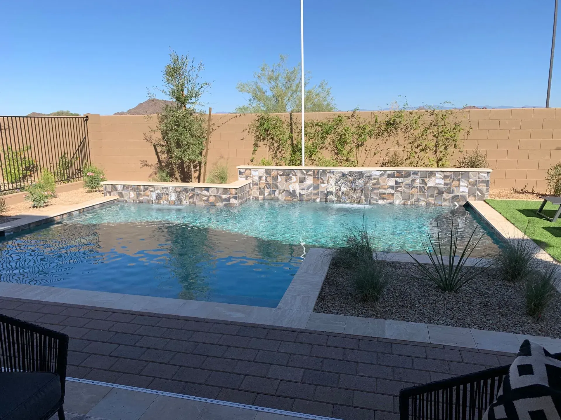 Pool with stone waterfall, surrounded by concrete, landscaping, and a brown wall, under a clear, blue sky.