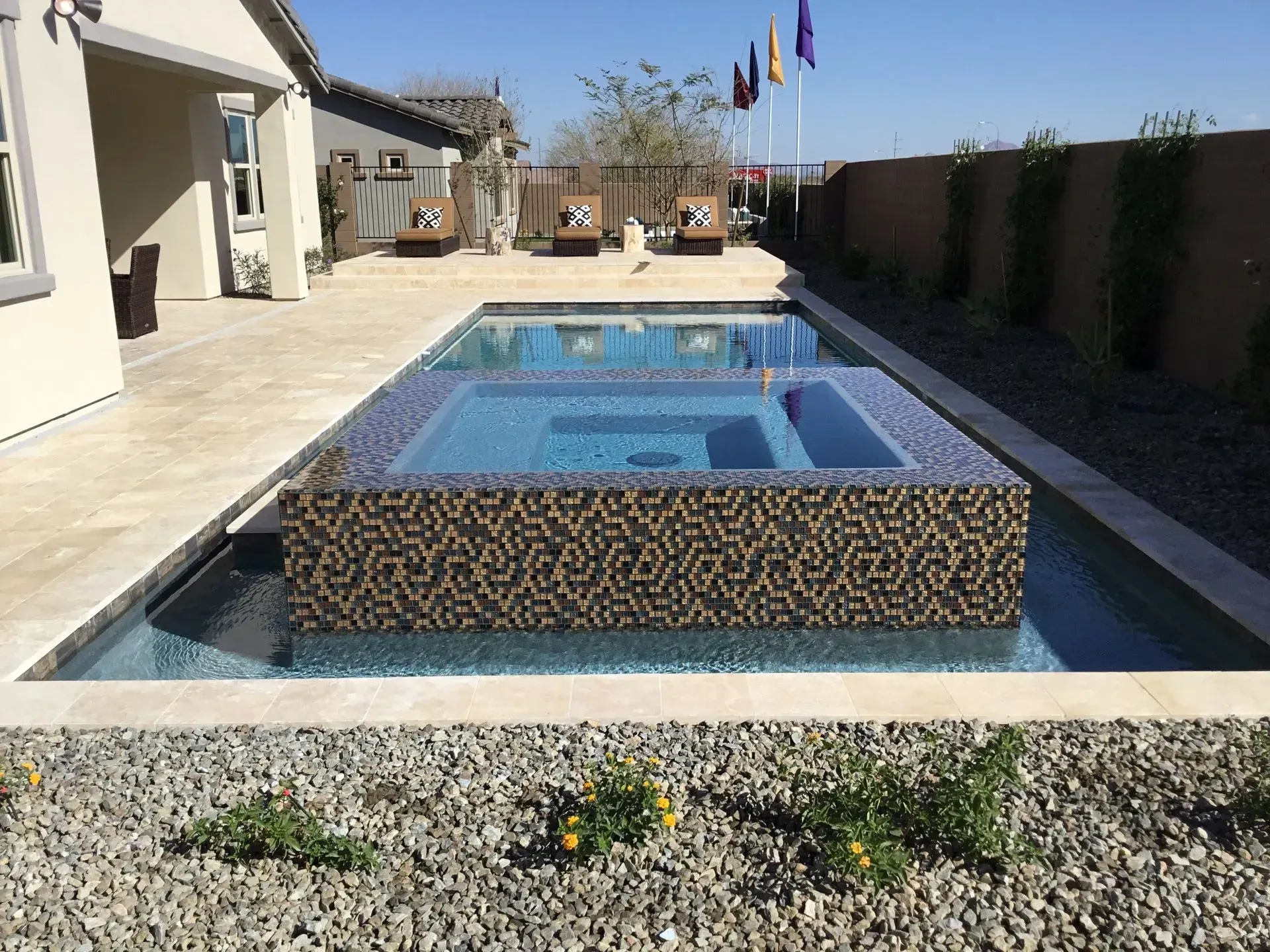Pool with raised spa; brown mosaic tiles; concrete patio; lounge chairs; blue sky.