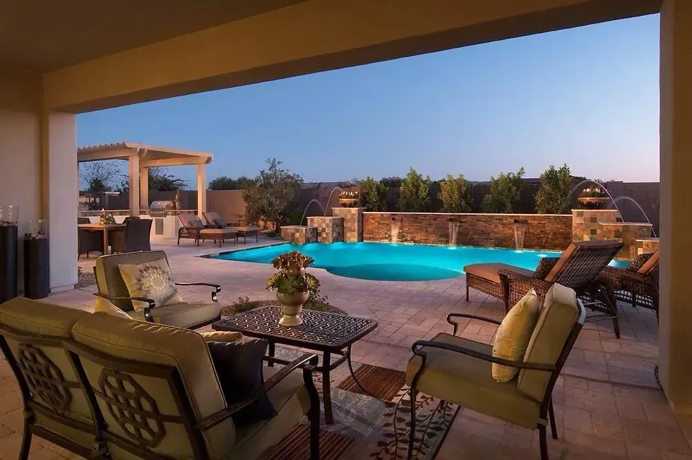 Patio with seating overlooking a pool, water fountains, and pergola at dusk.