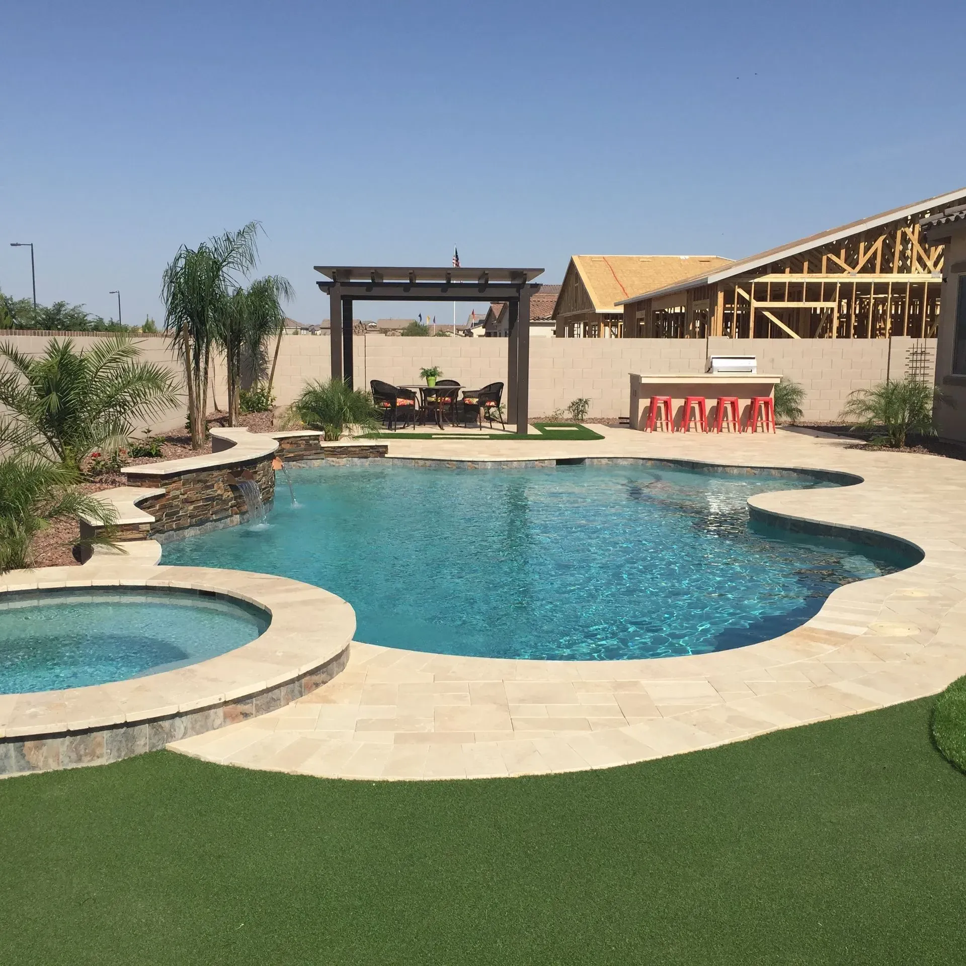 Swimming pool with spa, patio, and pergola in a backyard. House under construction in the background. Sunny day.