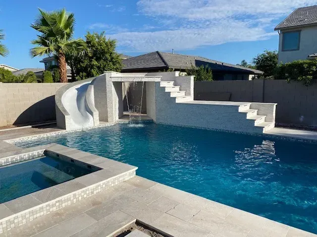 Swimming pool with water slide, stairs, spa, and palm tree under a blue sky.