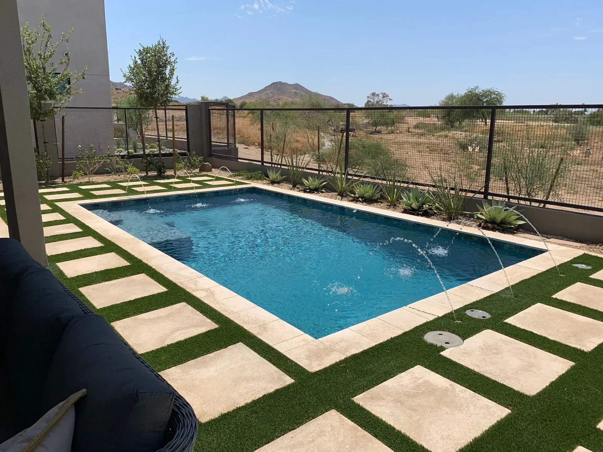 Rectangular pool with water features, surrounded by green turf and stone pavers. Fenced yard, desert landscape.