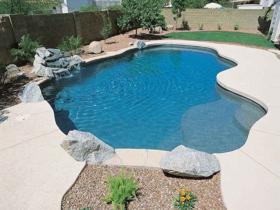 Swimming pool with blue water, surrounded by tan concrete and landscaping with rocks and plants.