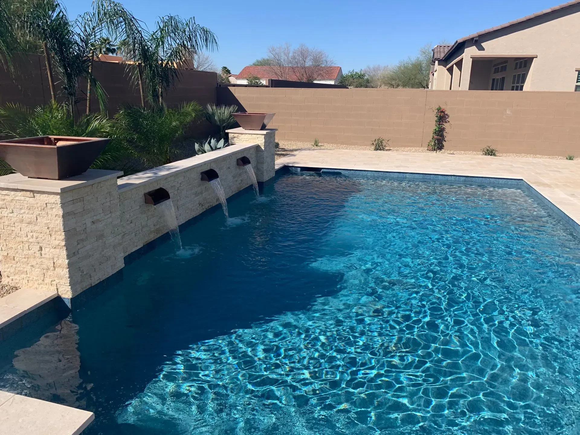 Swimming pool with water features, turquoise water, surrounded by tan stone and a brown wall.