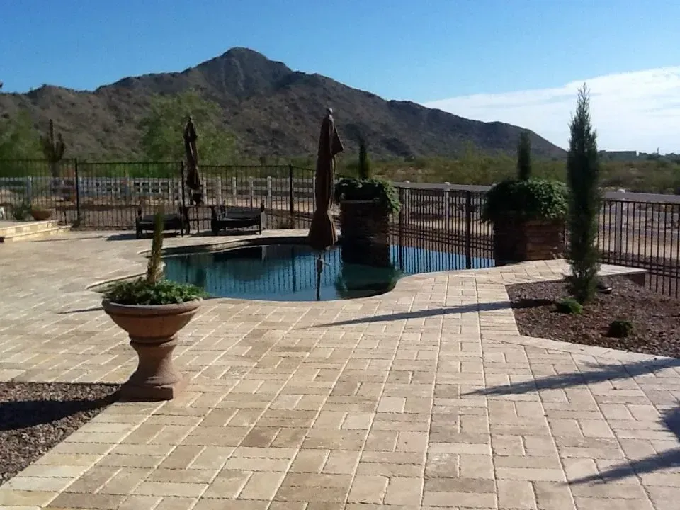 Pool and patio with mountain backdrop; sunny day. Beige pavers, dark pool, black fence, potted plants.