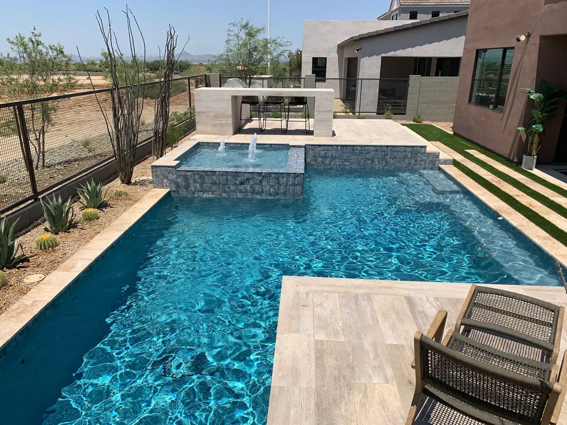 A modern backyard with a pool, spa, and outdoor kitchen. Blue water, gray tile, beige and brown building.