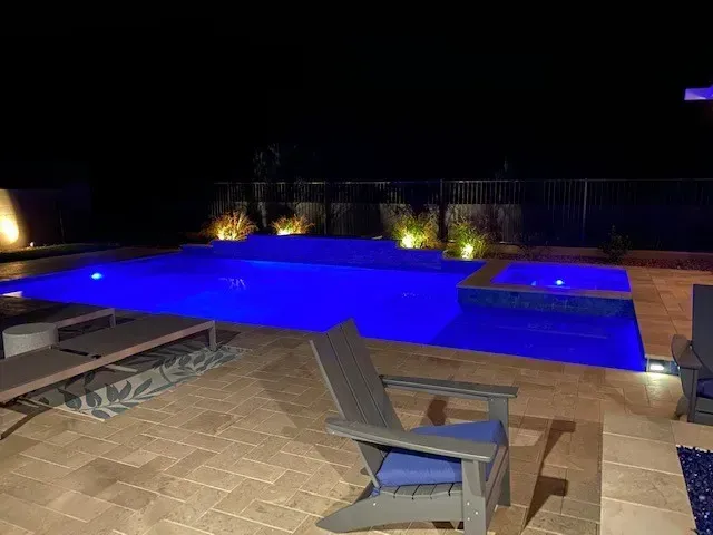 Nighttime view of a blue-lit swimming pool and spa. Patio furniture surrounds the pool. Dark background with uplighting.