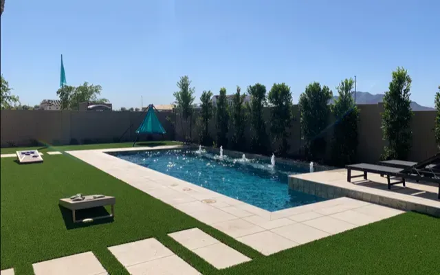 Backyard with pool, fountains, artificial grass, lounge chairs, and cornhole game under a clear blue sky.