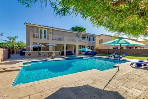 Backyard with pool, patio, and two-story house. Blue pool, beige patio, lounge chairs, and umbrellas.