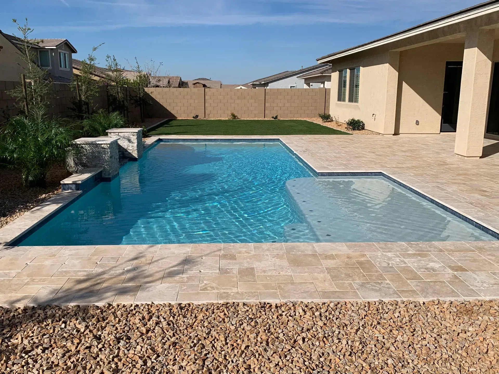 Pool with turquoise water surrounded by beige tiles and gravel, next to a beige house under a blue sky.
