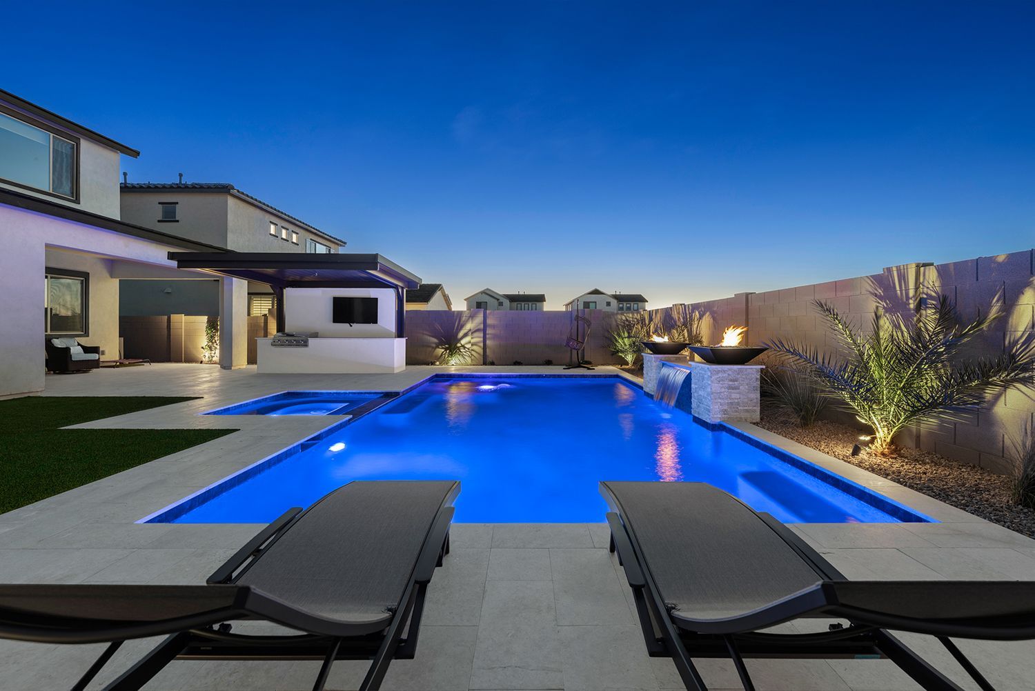 Backyard pool at dusk with blue illuminated water, lounge chairs, and a fire pit.