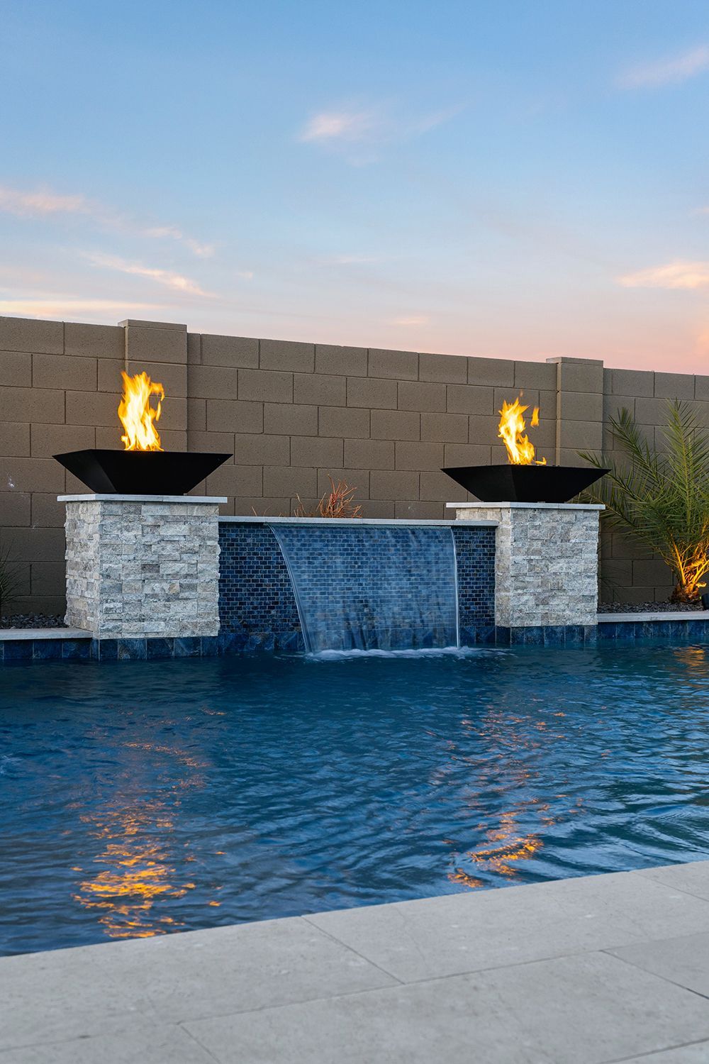 Pool with waterfall feature, fire bowls, and textured stone wall, at sunset.