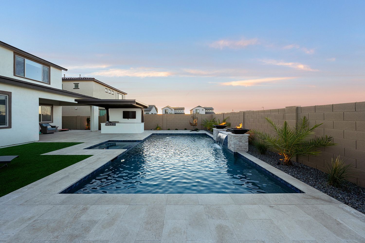 Backyard pool with a waterfall feature, patio, and modern house under a sunset sky.