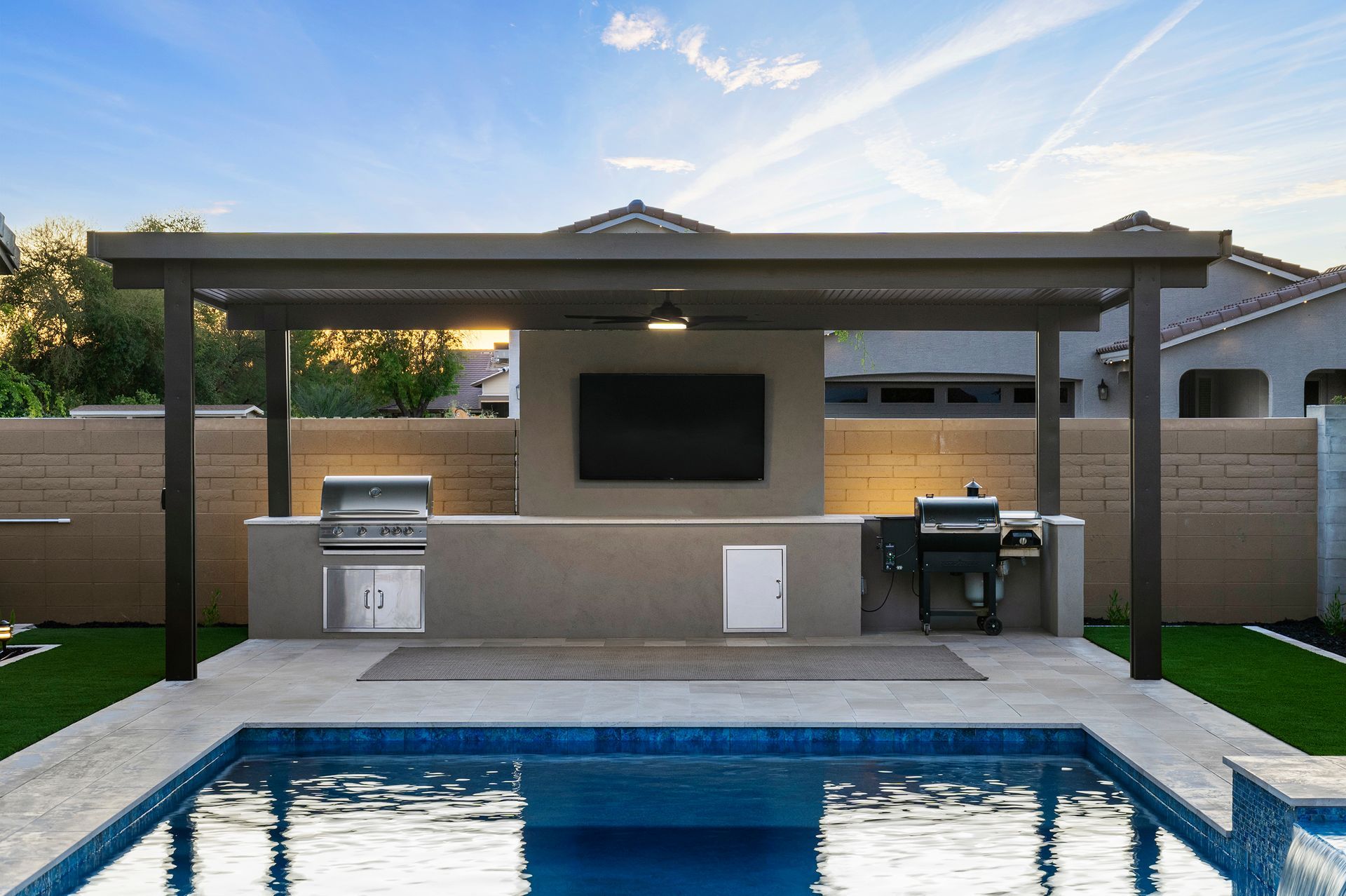 Poolside outdoor kitchen with a pergola, grilling area, TV, and pool.