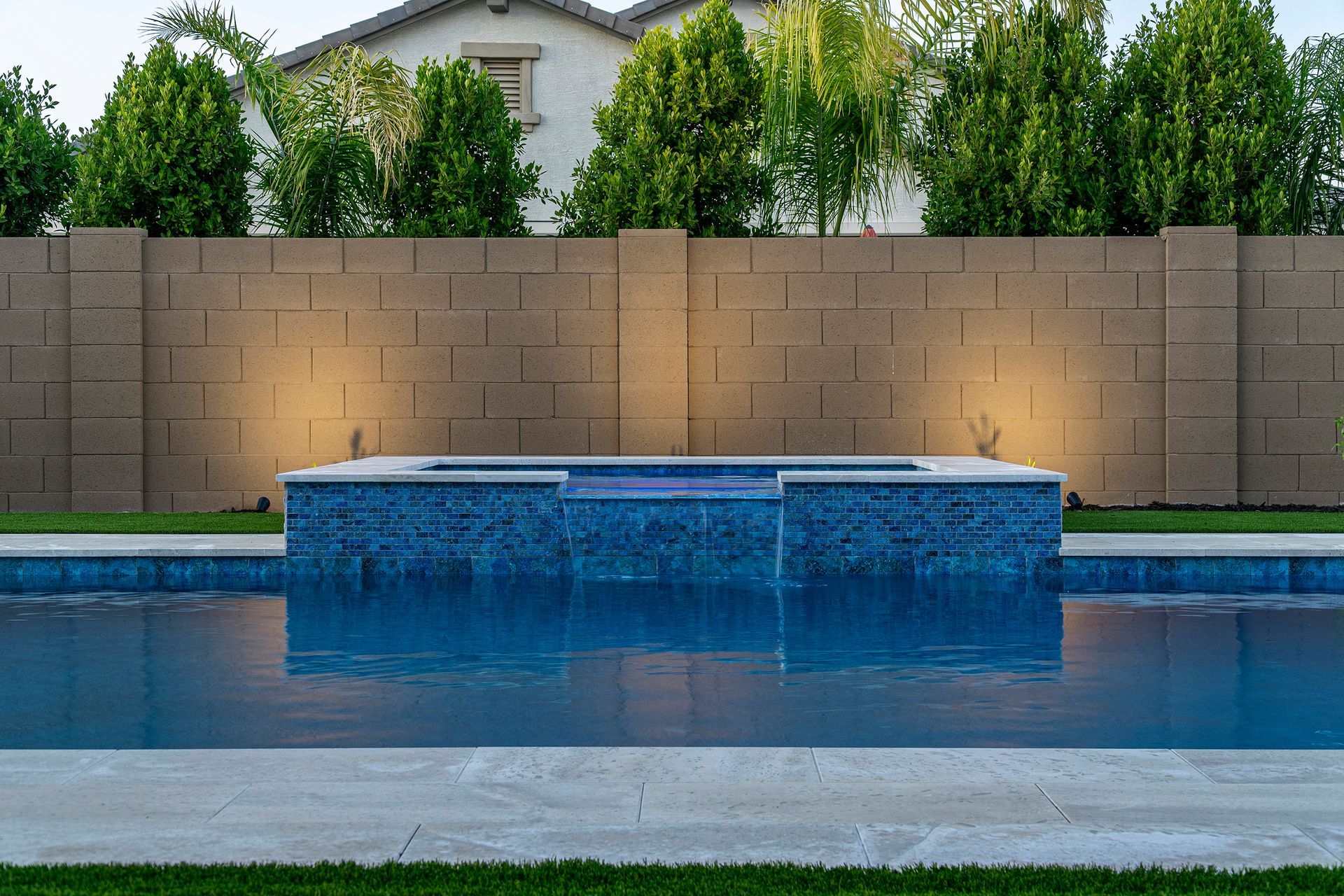 Blue tiled pool with a raised spa, against a tan brick wall with green trees.