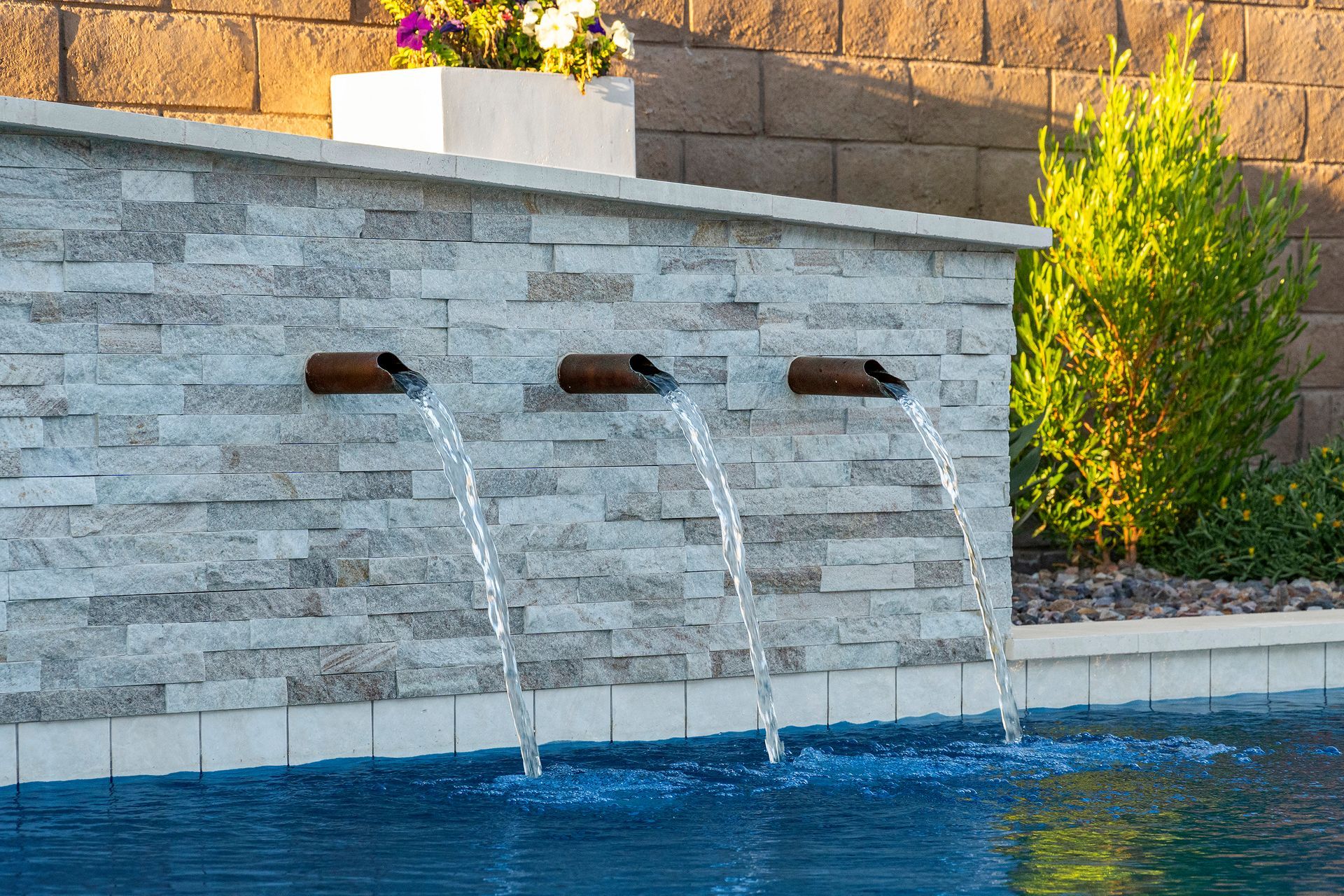 Water fountain with three spouts cascading into a pool, against a stone wall and brick background.